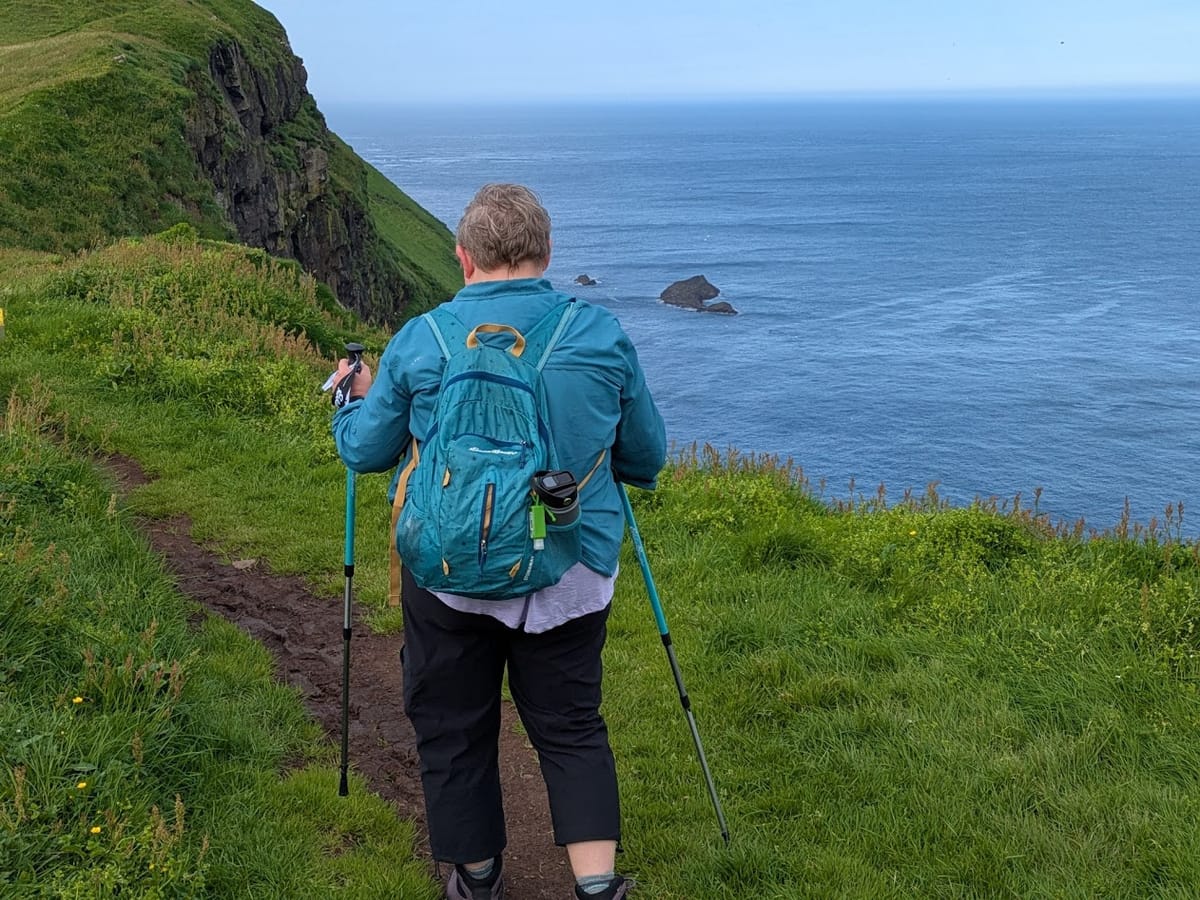 A woman pictured from behind, hiking along a pathway that overlooks the ocean. She is using turquoise hiking poles and is wearing a teal backpack.