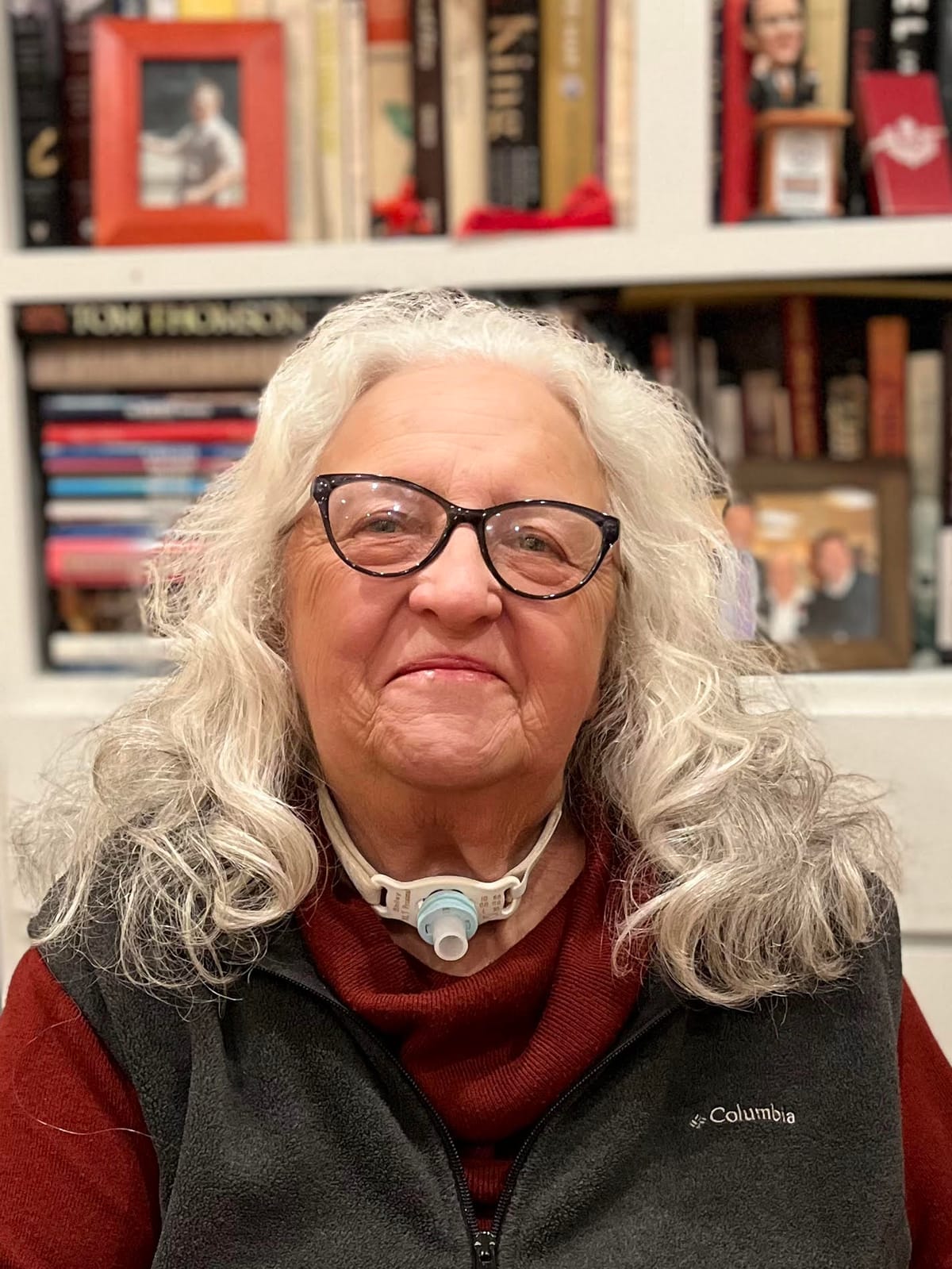 An woman with shoulder-length wavy gray hair smiles at the camera. She is wearing glasses, a rust coloured turtleneck and a dark gray fleece vest. She sits in front of a bookcase. She has