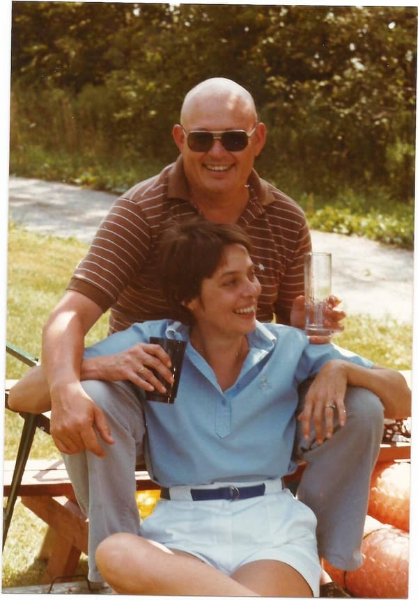 A couple sits casually together. He is bald, wearing sunglesses. She has brown hair, is smiling and has her arms draped over his legs. Both are holding empty glasses