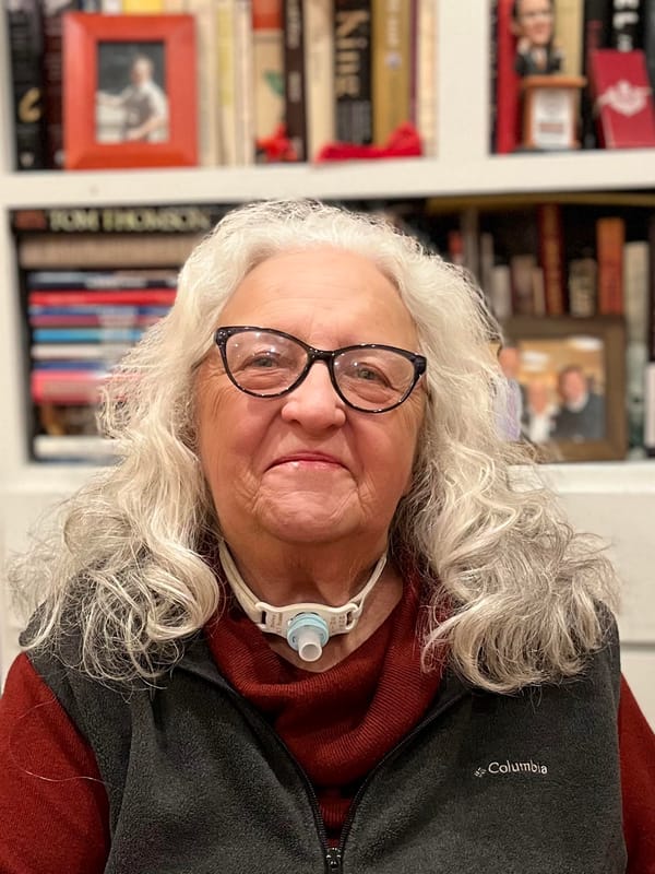 An woman with shoulder-length wavy gray hair smiles at the camera. She is wearing glasses, a rust coloured turtleneck and a dark gray fleece vest.  She sits in front of a bookcase. She has
